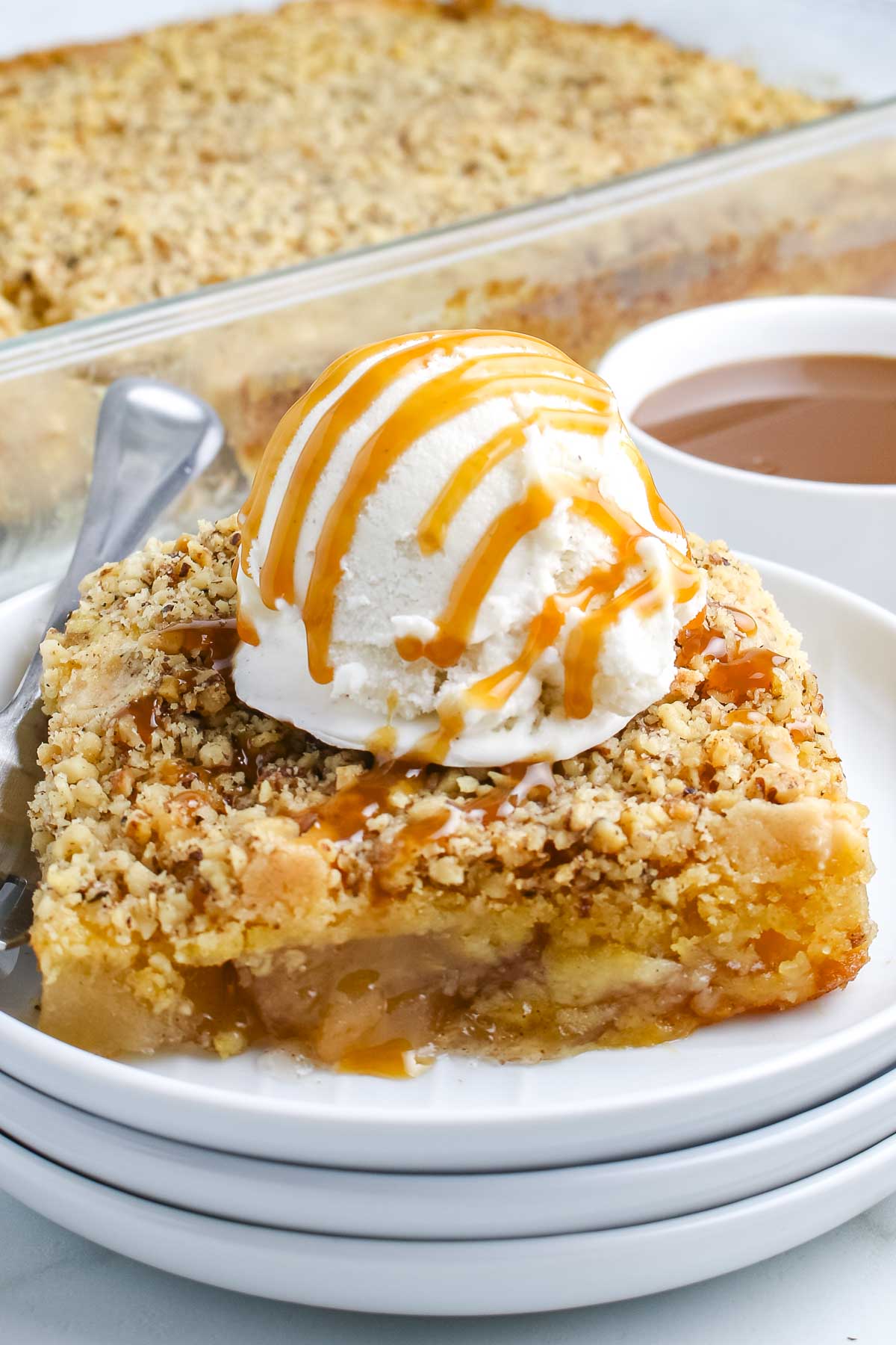 A close-up view of a square slice of warm apple dump cake topped with a scoop of vanilla ice cream and a drizzle of caramel sauce on a white plate.