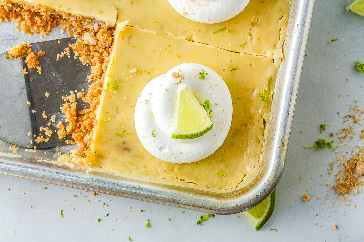 A close-up view of a partially sliced key lime pie in a metal baking pan with a metal serving spatula resting inside. The pie is topped with whipped cream and small pieces of lime.