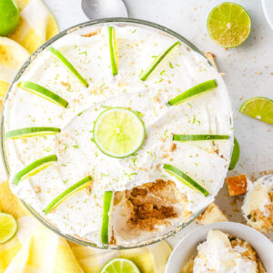 An overhead view of a large key lime trifle in a glass bowl sitting on a yellow checkered napkin. The top features whipped cream and lime slices arranged in a circle with a large portion scooped out to reveal the interior layers.