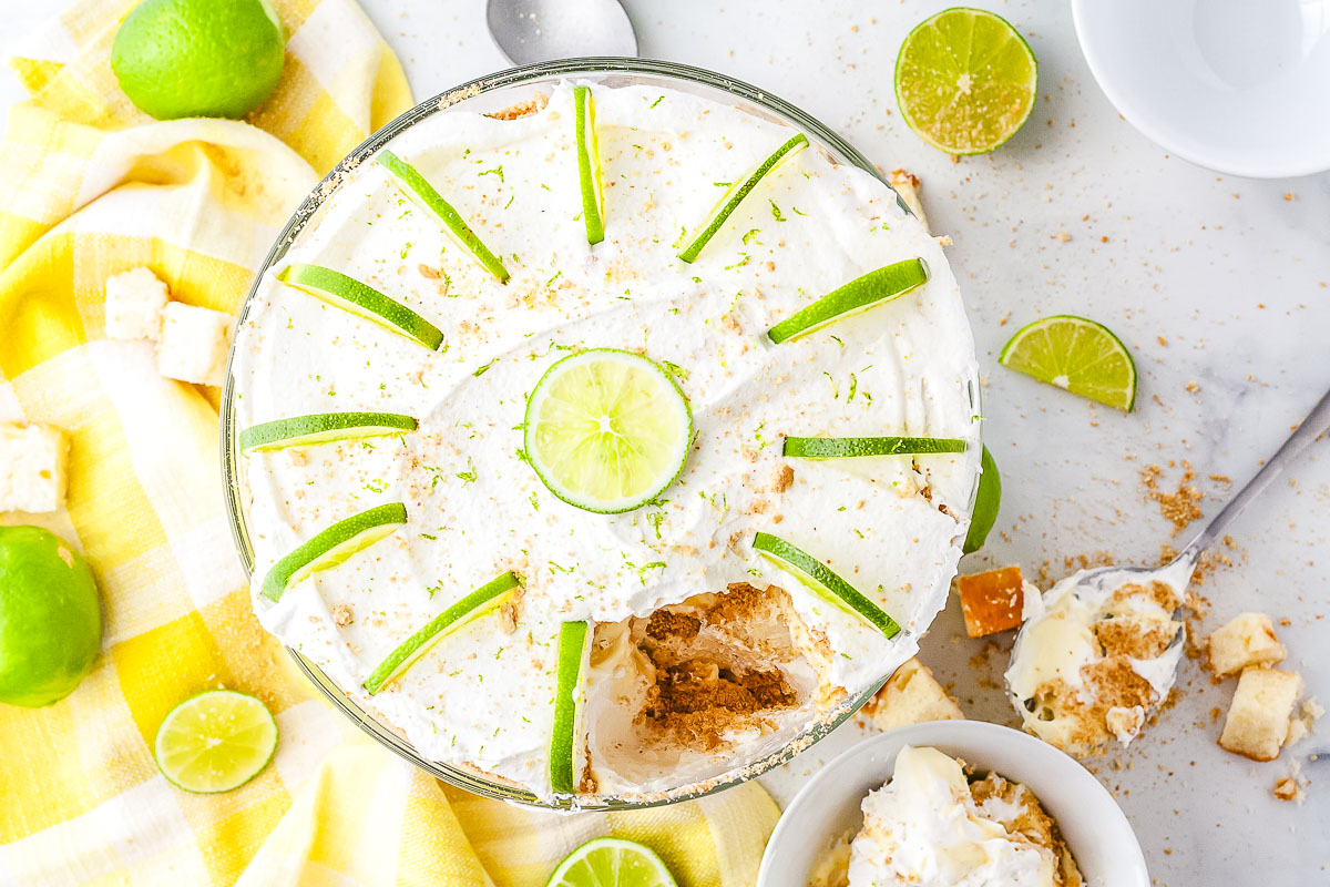 An overhead view of a large key lime trifle in a glass bowl sitting on a yellow checkered napkin. The top features whipped cream and lime slices arranged in a circle with a large portion scooped out to reveal the interior layers.