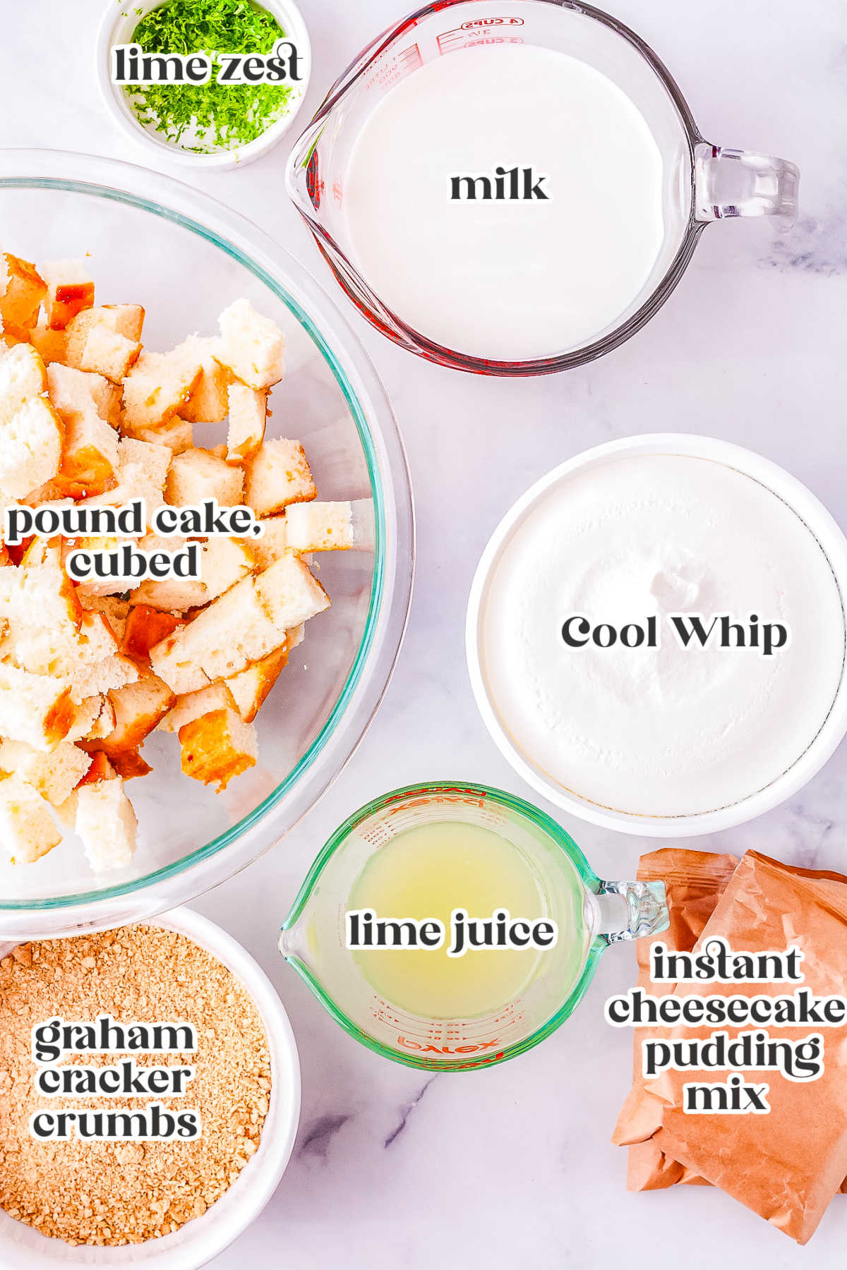 A flat lay showing labeled ingredients needed to make a key lime trifle. The items include cubed pound cake, graham cracker crumbs, fresh lime juice, instant cheesecake pudding mix, Cool Whip, cold milk, and lime zest.