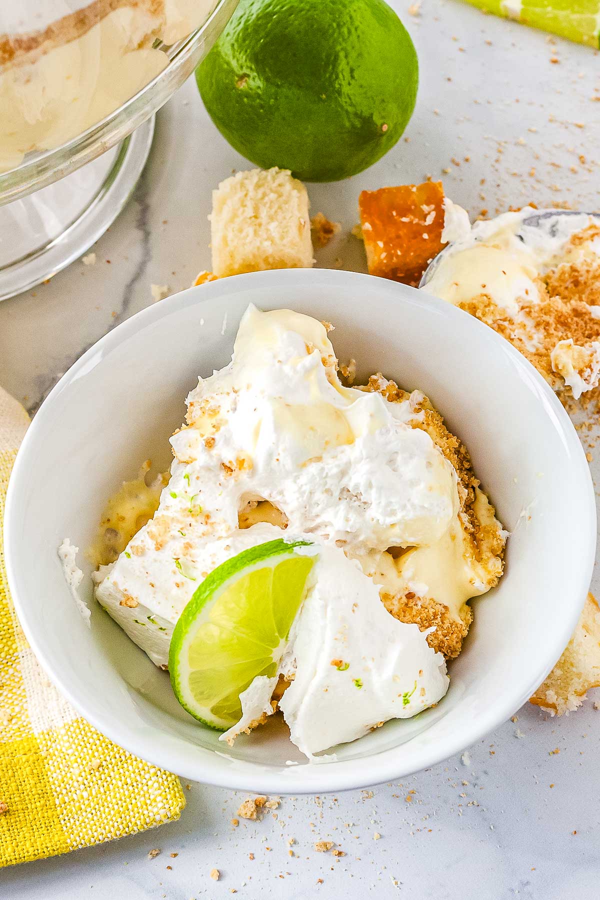 A white serving bowl filled with a scooped portion of layered key lime trifle. The dessert shows pieces of pound cake, yellow filling, graham cracker crumbs, and whipped cream garnished with a fresh lime slice.