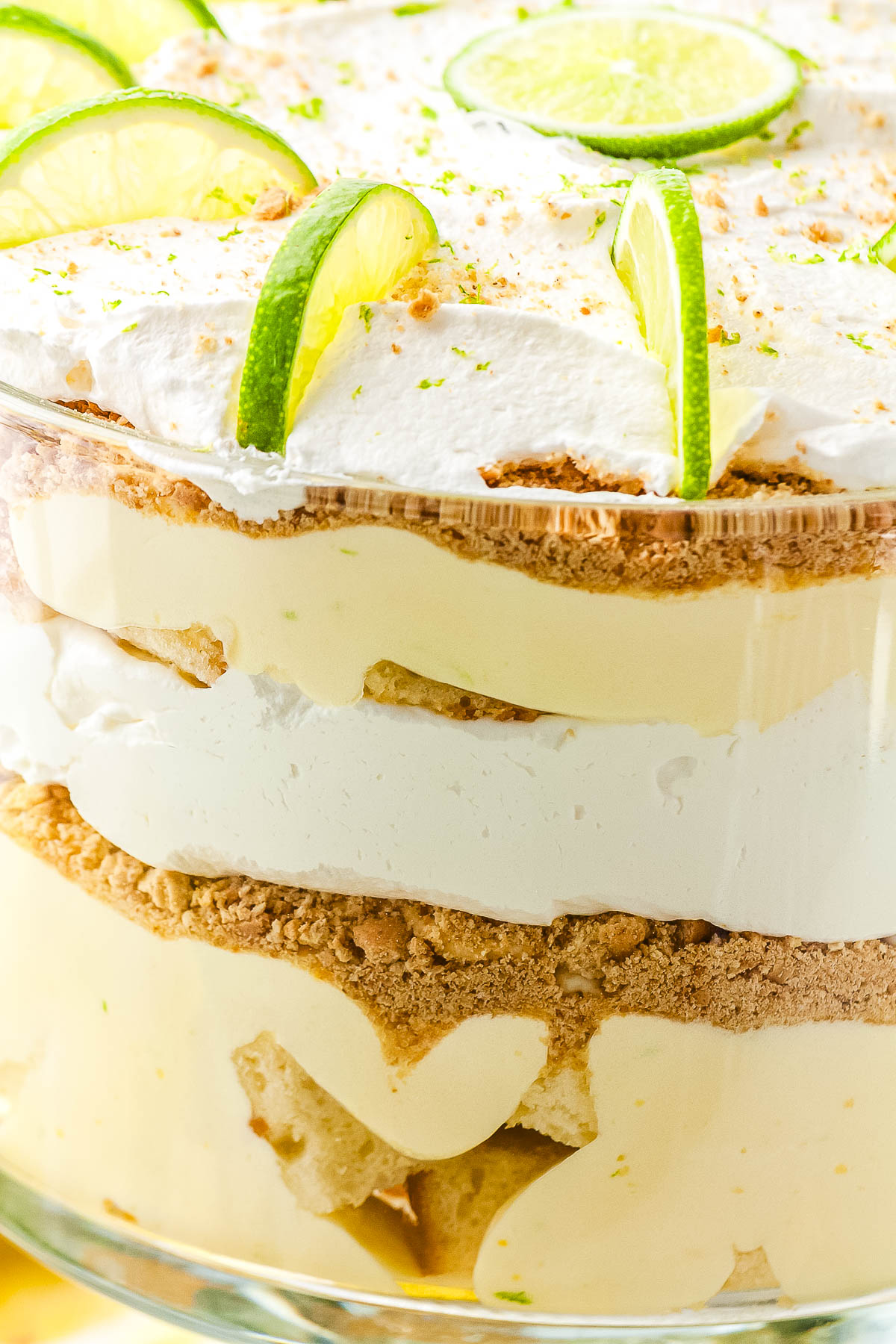 A close-up side view of a glass trifle bowl filled with a key lime dessert. The clear sides show the distinct layers of cubed pound cake, yellow lime filling, crushed graham crackers, and white whipped cream.