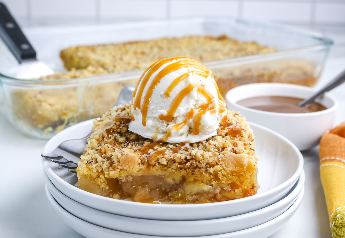 A plated slice of apple dump cake topped with vanilla ice cream and caramel sauce sits in front of a glass baking dish filled with the remaining dessert.