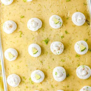 An overhead view of a large sheet pan key lime pie in a metal pan. The dessert is covered with evenly spaced dollops of whipped cream and scattered green lime zest.