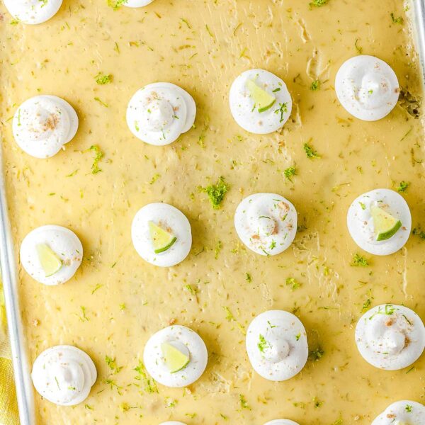 An overhead view of a large sheet pan key lime pie in a metal pan. The dessert is covered with evenly spaced dollops of whipped cream and scattered green lime zest.