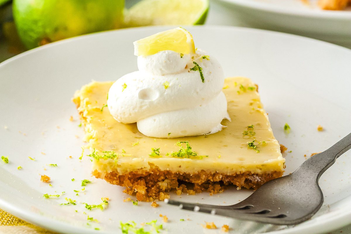 A close-up view of a square slice of key lime pie resting on a white plate next to a metal fork. The dessert features a graham cracker crust and a smooth lime filling topped with whipped cream.