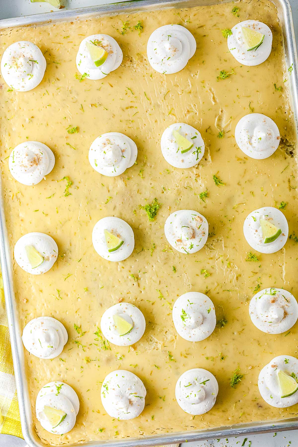 An overhead view of a large sheet pan key lime pie in a metal pan. The dessert is covered with evenly spaced dollops of whipped cream and scattered green lime zest.