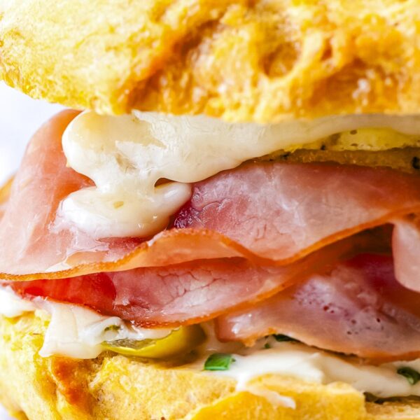 A close-up side view of a breakfast bagel sandwich showing layers of honey ham, melted cheese, sausage, and jalapeños between a toasted egg bagel.