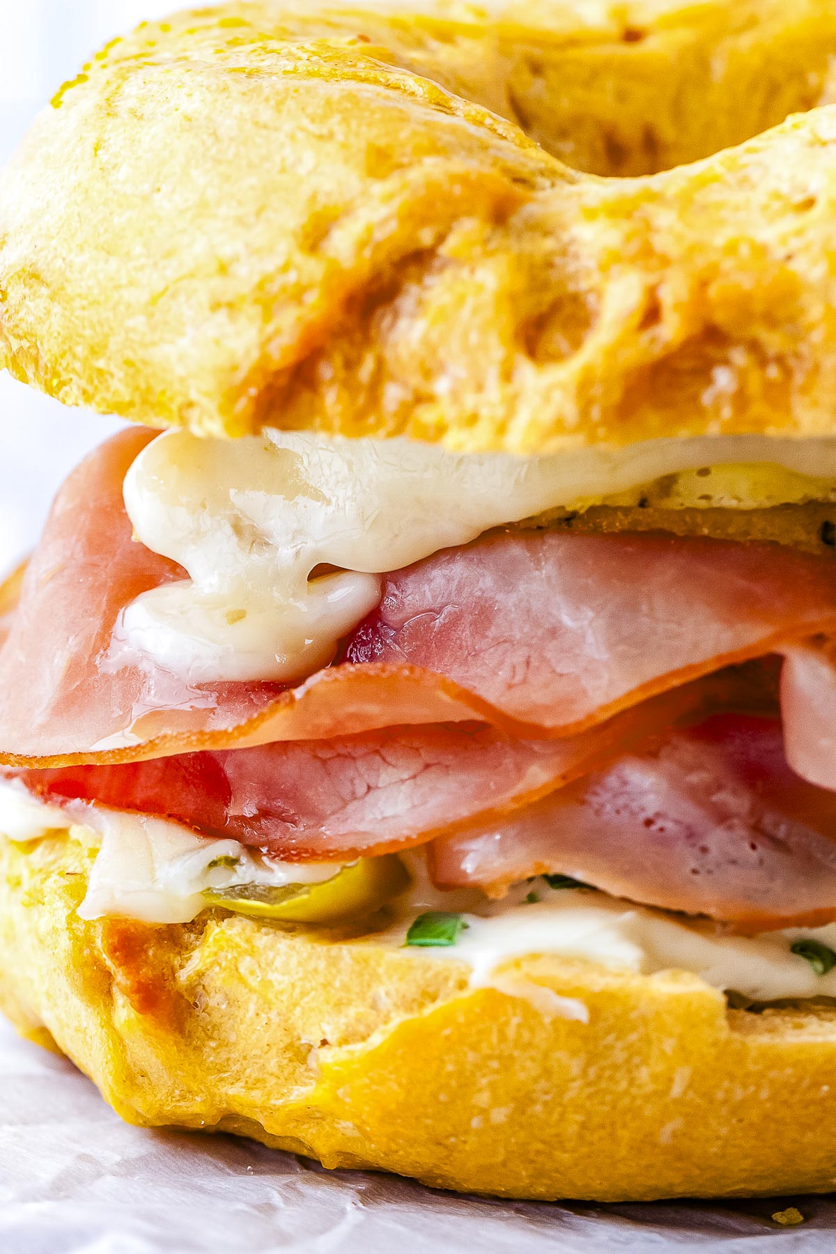 A close-up side view of a breakfast bagel sandwich showing layers of honey ham, melted cheese, sausage, and jalapeños between a toasted egg bagel.
