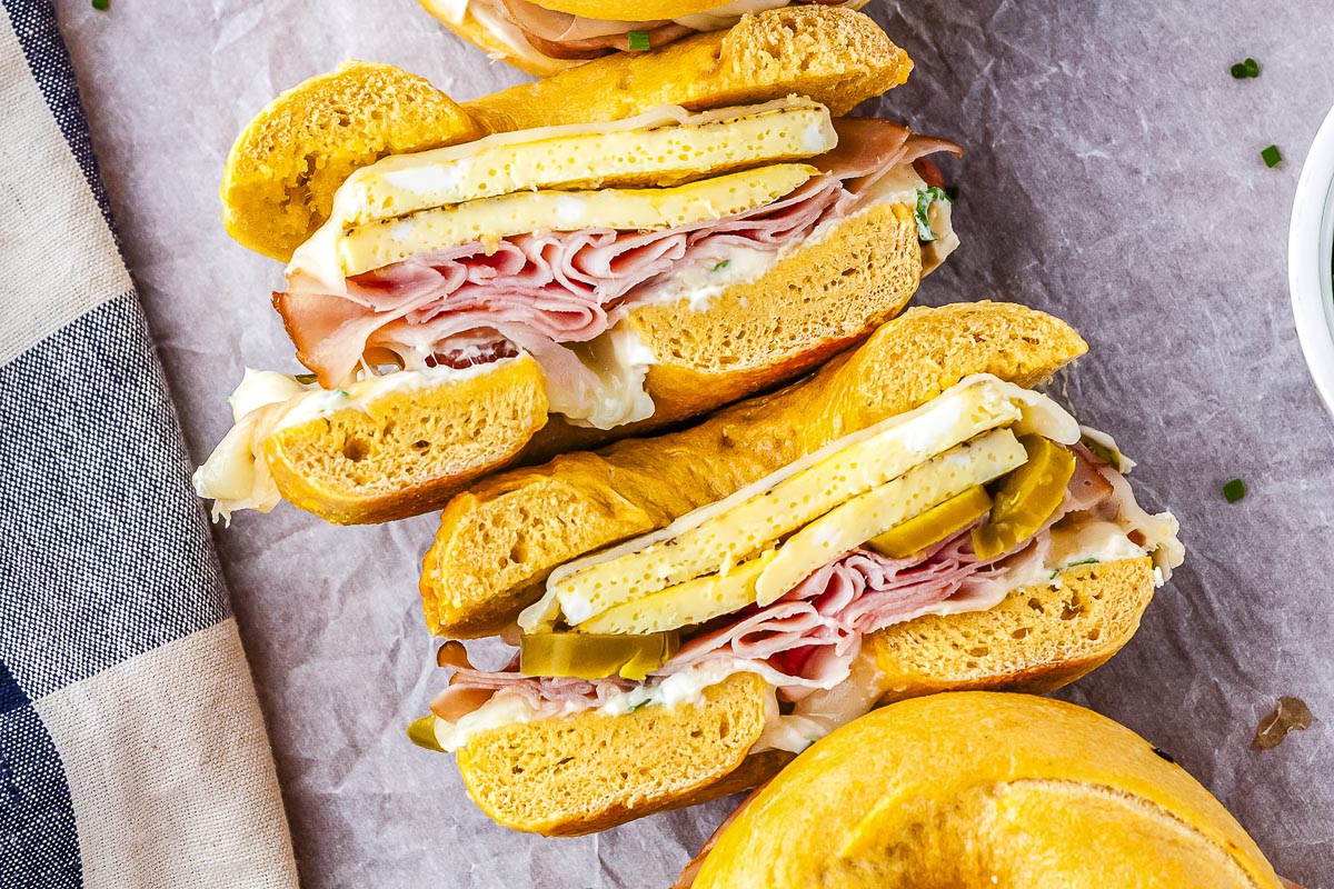 An overhead view of several breakfast bagel sandwiches sliced in half, showing the layers of egg, ham, sausage, and cream cheese stacked together on parchment paper.