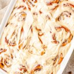 An overhead view of a white rectangular baking dish filled with baked apple cinnamon rolls. The warm rolls are completely covered in a thick layer of melted cream cheese icing.