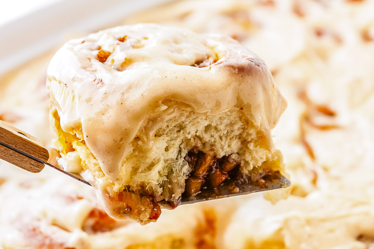 A close-up view of a frosted apple pie cinnamon roll lifted on a metal spatula. The side of the warm roll reveals layers of fluffy dough filled with diced apples and cinnamon sugar.