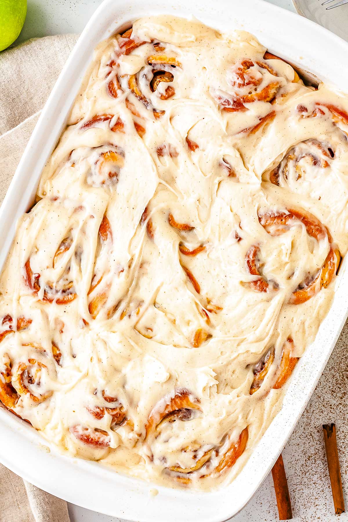 An overhead view of a white rectangular baking dish filled with baked apple cinnamon rolls. The warm rolls are completely covered in a thick layer of melted cream cheese icing.