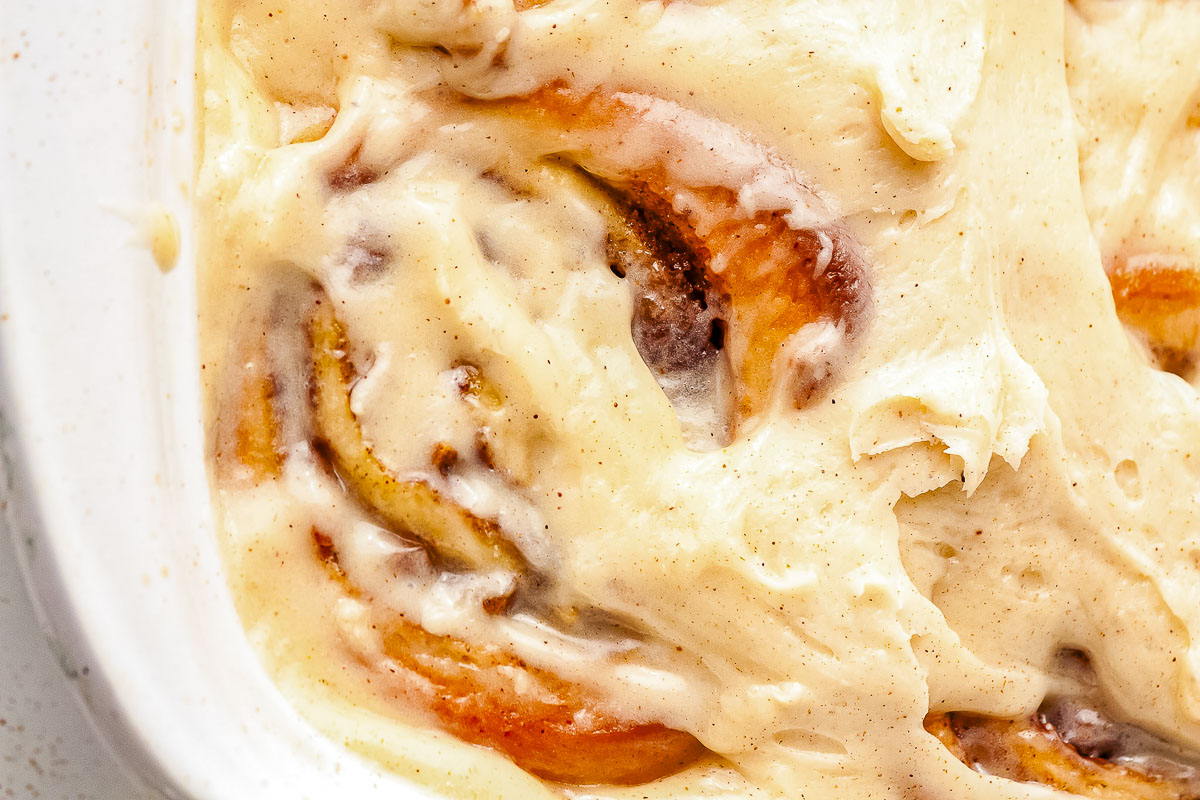 An extreme close-up view of baked apple pie cinnamon rolls in a pan. Thick cream cheese icing melts into the spiral folds of the dough, revealing hints of cinnamon and cooked apples.