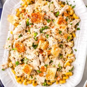 An overhead view of a large white serving platter filled with chicken pot pie pasta. The creamy dish is garnished with square puff pastry croutons, Parmesan cheese, and fresh parsley, resting next to a blue and white checkered napkin.