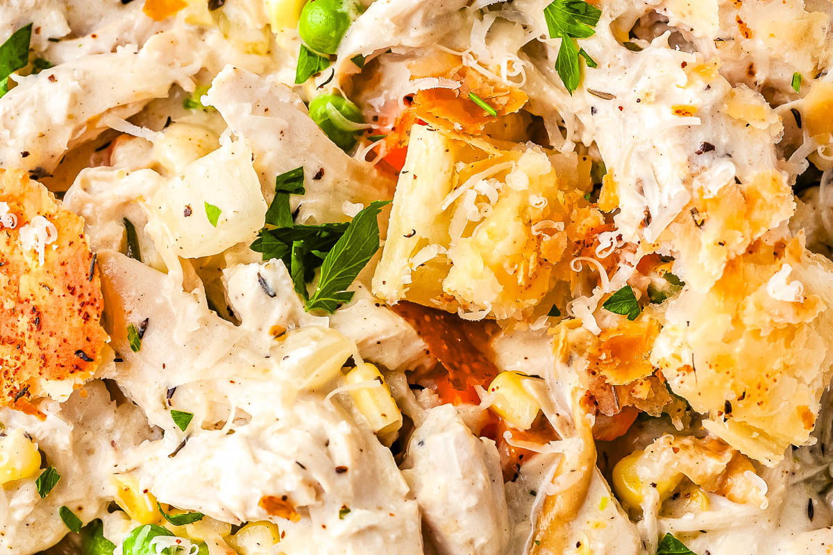 A close-up view of creamy chicken pot pie pasta showing shredded chicken, cavatappi noodles, peas, and corn covered in a rich cream sauce. The dish is topped with flaky puff pastry croutons, grated Parmesan, and fresh parsley.