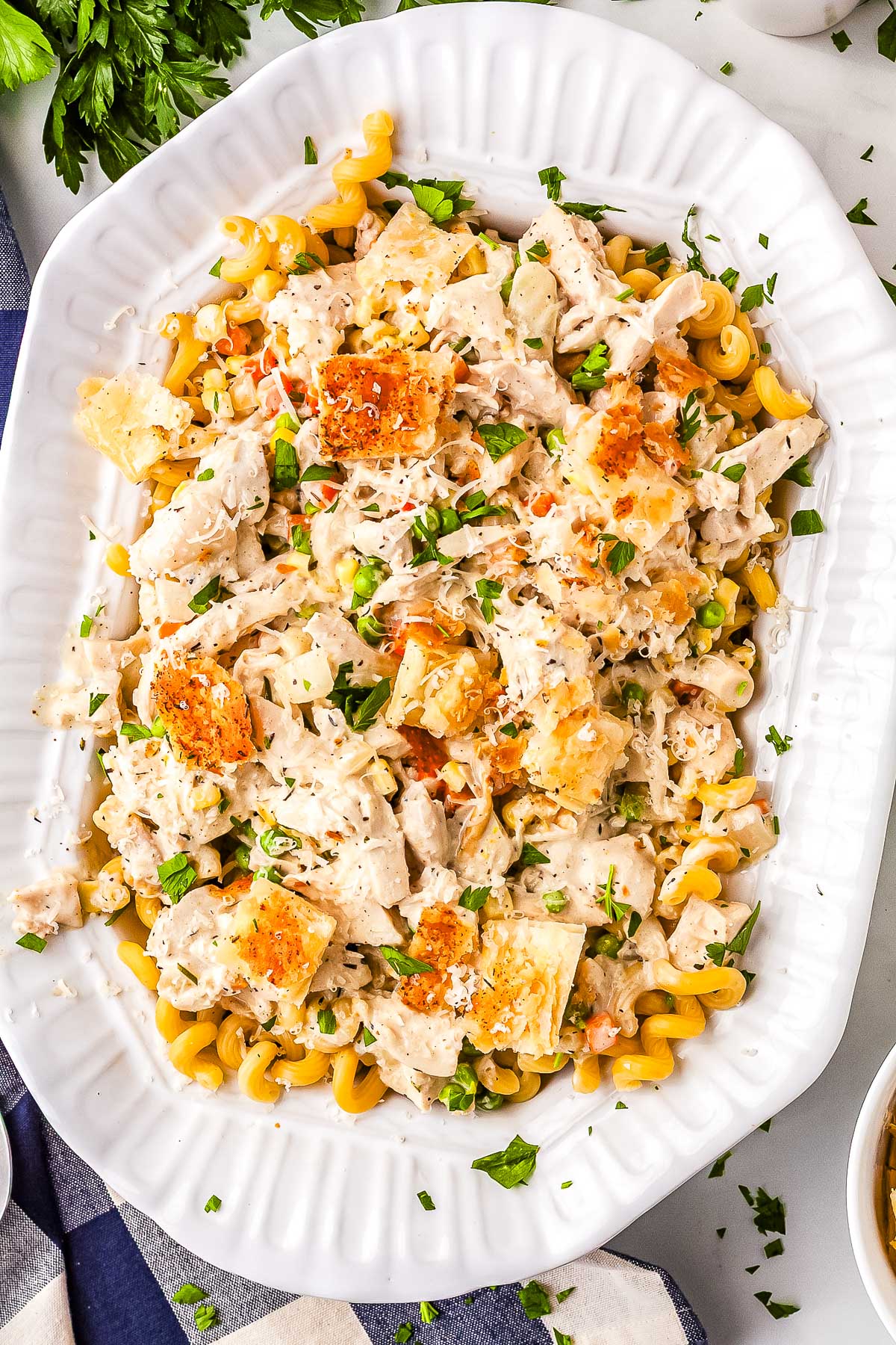 An overhead view of a large white serving platter filled with chicken pot pie pasta. The creamy dish is garnished with square puff pastry croutons, Parmesan cheese, and fresh parsley, resting next to a blue and white checkered napkin.