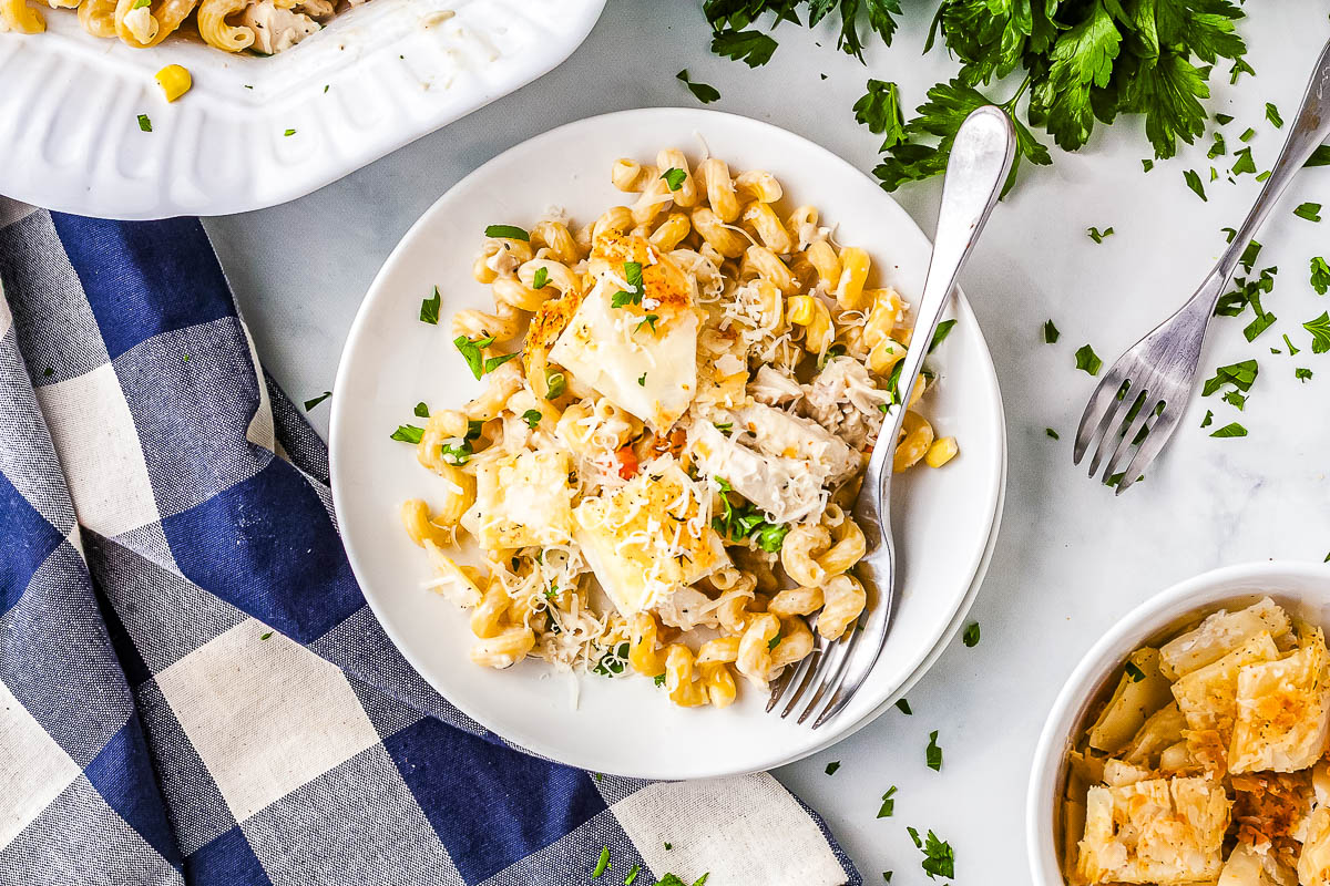 A white plate holding a portion of creamy chicken pot pie pasta topped with puff pastry croutons and fresh parsley. A silver fork rests on the edge of the plate over a blue and white checkered napkin.