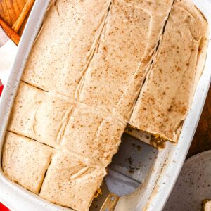 An overhead view of a frosted olive oil pumpkin cake sliced into squares inside a white rectangular baking pan. A metal serving spatula rests inside the pan where a slice has been removed.