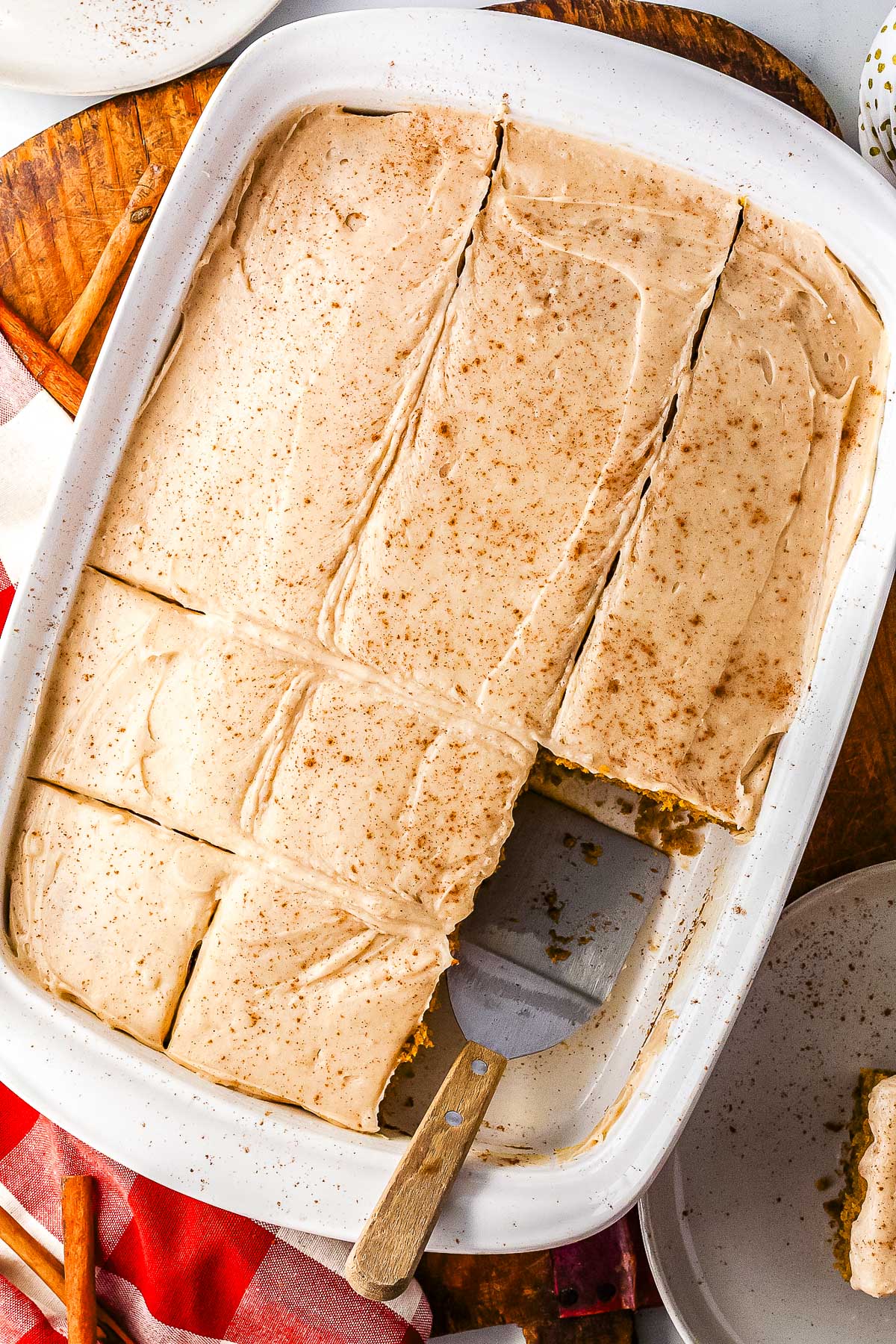 An overhead view of a frosted olive oil pumpkin cake sliced into squares inside a white rectangular baking pan. A metal serving spatula rests inside the pan where a slice has been removed.