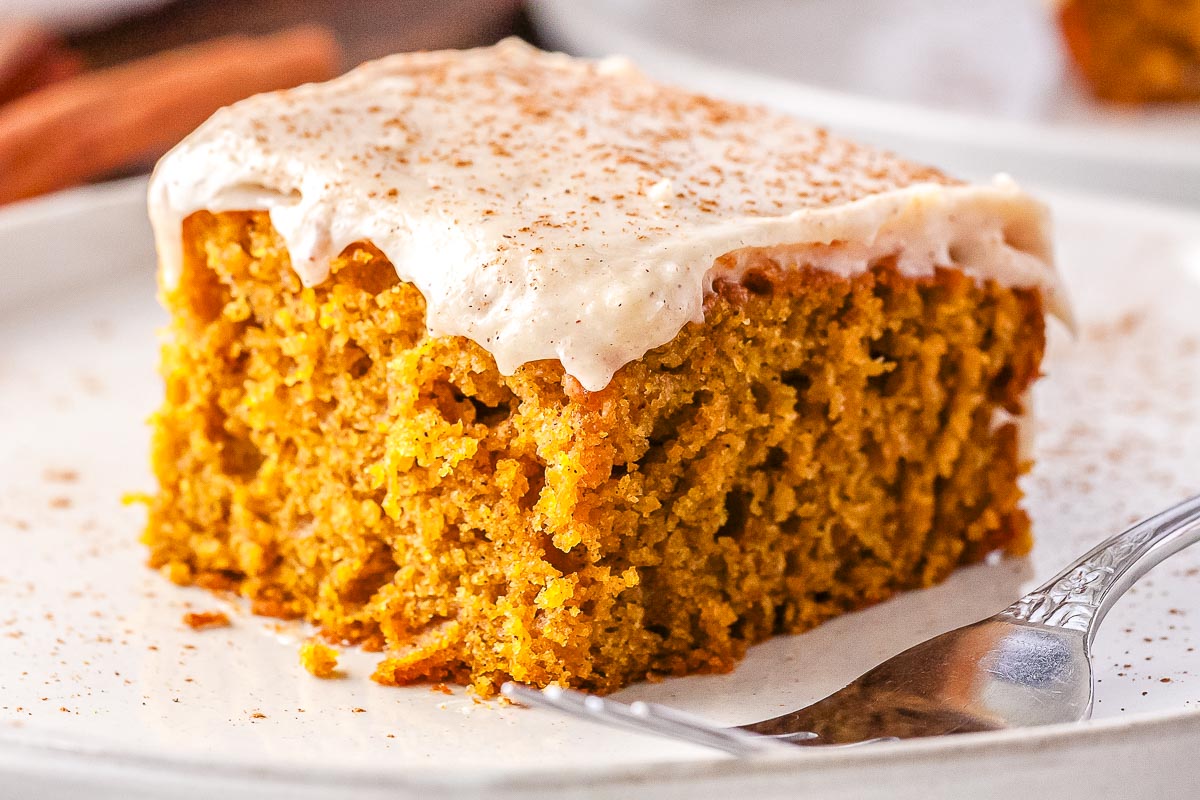 A close-up view of a square slice of pumpkin olive oil cake on a white plate. The moist orange sponge is topped with a thick layer of cream cheese frosting and a light sprinkle of cinnamon.