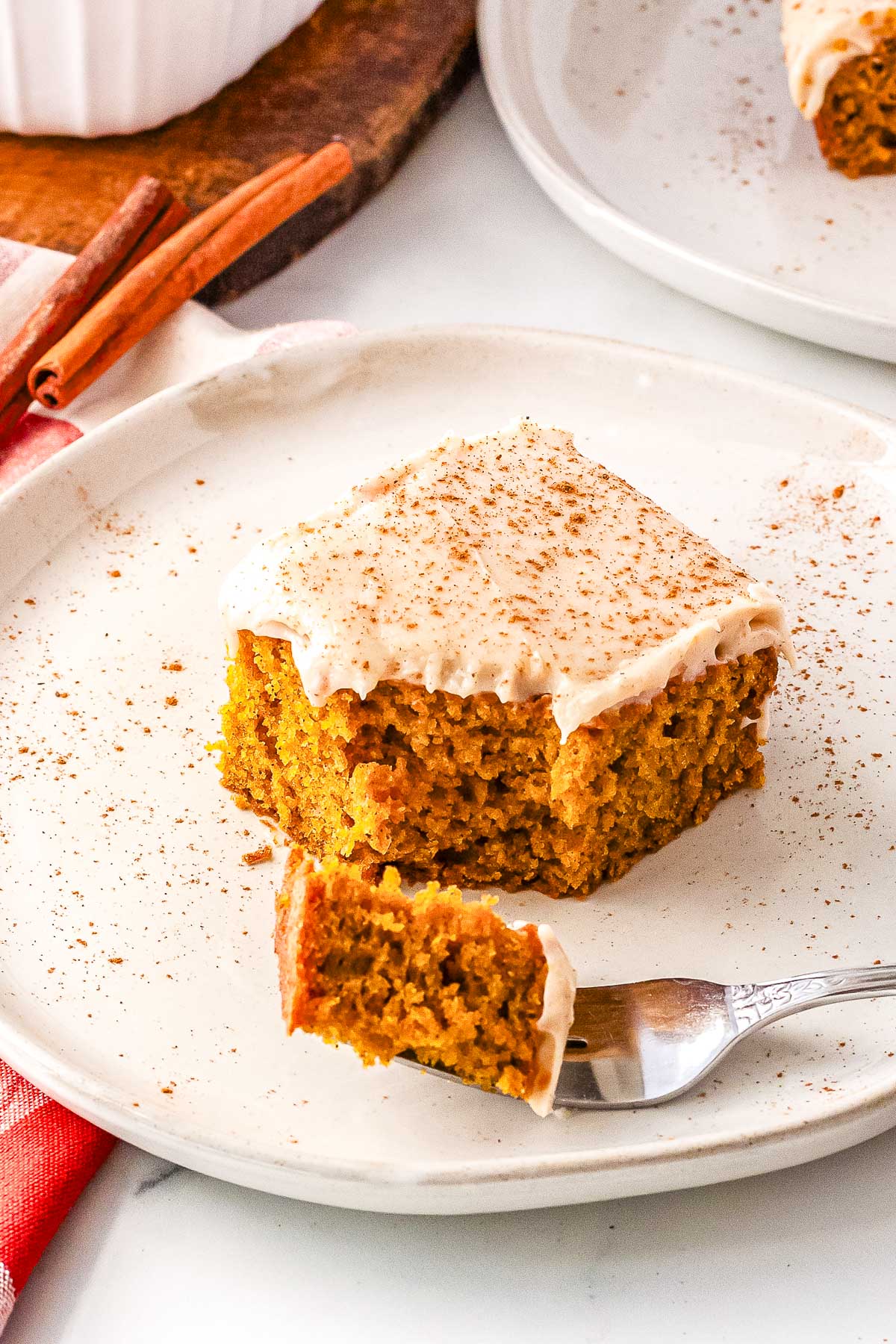 A white plate holding a slice of frosted pumpkin olive oil cake with a bite taken out of it. A silver fork holds the bite-sized piece of cake in the foreground, next to a red checkered napkin.