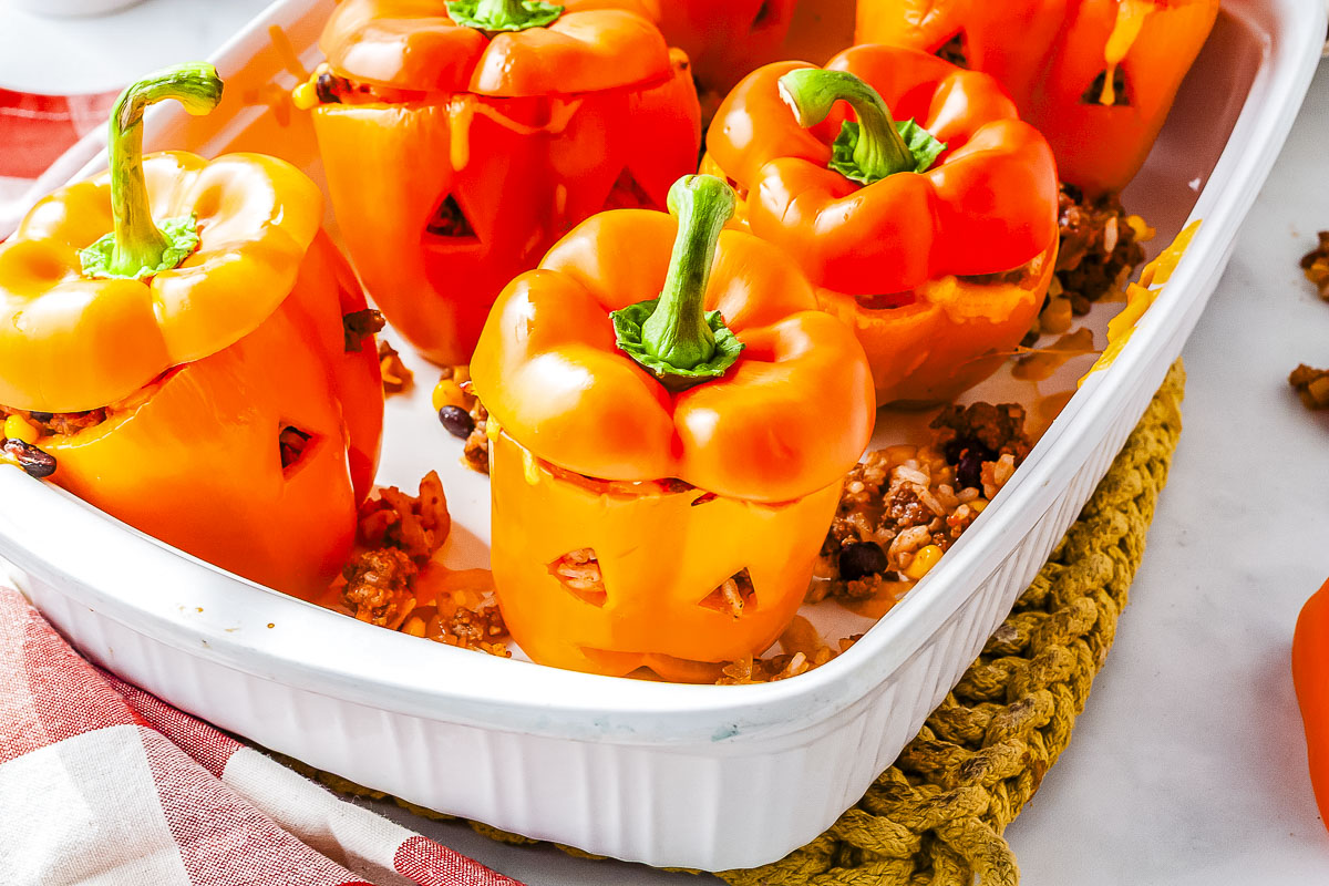 An angled view of six baked orange bell peppers carved with jack-o'-lantern faces, filled with seasoned meat and rice, arranged inside a white 9x13-inch baking dish.