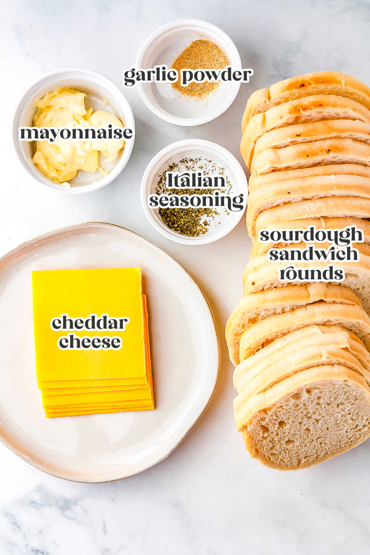 An overhead flat lay of ingredients labeled for Halloween grilled cheese, including sourdough sandwich rounds, sliced cheddar cheese, mayonnaise, garlic powder, and Italian seasoning.