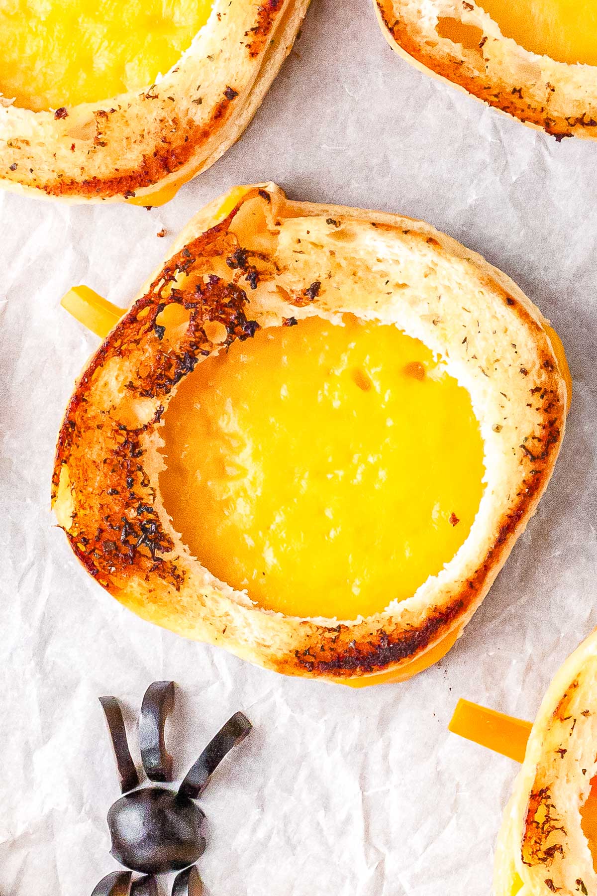 A close-up view of a golden brown pumpkin-shaped grilled cheese sandwich with melted cheddar cheese in the center and a black olive spider on white parchment paper.