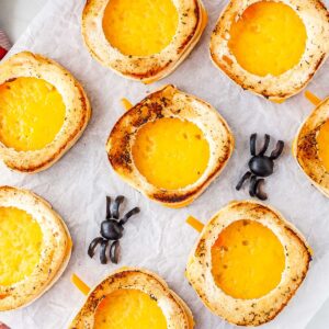 A platter of golden brown pumpkin-shaped grilled cheese sandwiches with bell pepper stems and black olive spiders resting on parchment paper.
