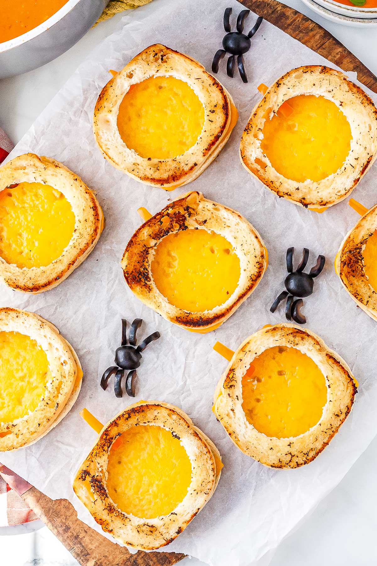 A platter of golden brown pumpkin-shaped grilled cheese sandwiches with bell pepper stems and black olive spiders resting on parchment paper.