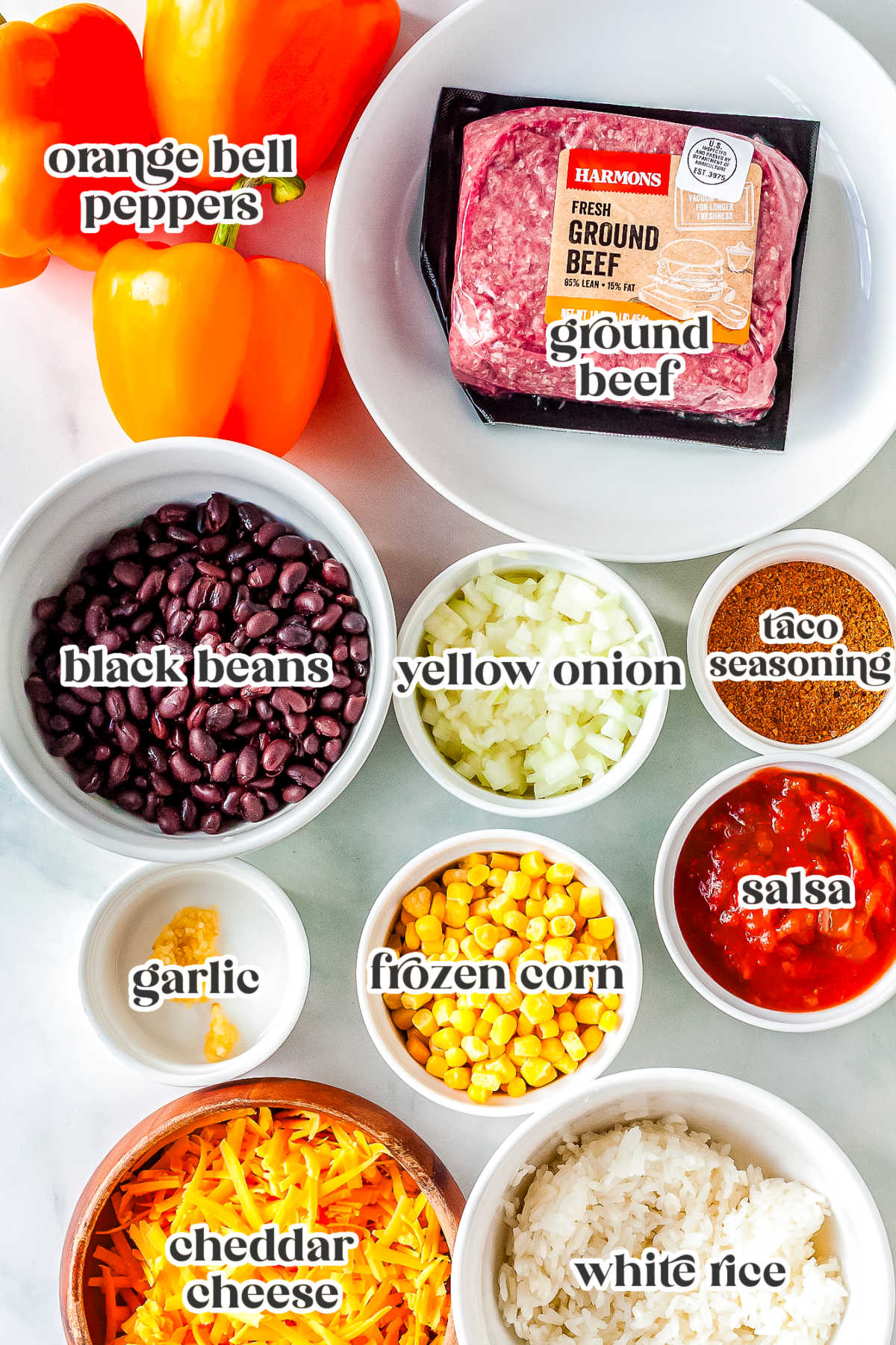 An overhead flat lay of recipe ingredients labeled in white bowls, including ground beef, orange bell peppers, black beans, yellow onion, taco seasoning, garlic, frozen corn, salsa, shredded cheddar cheese, and white rice.