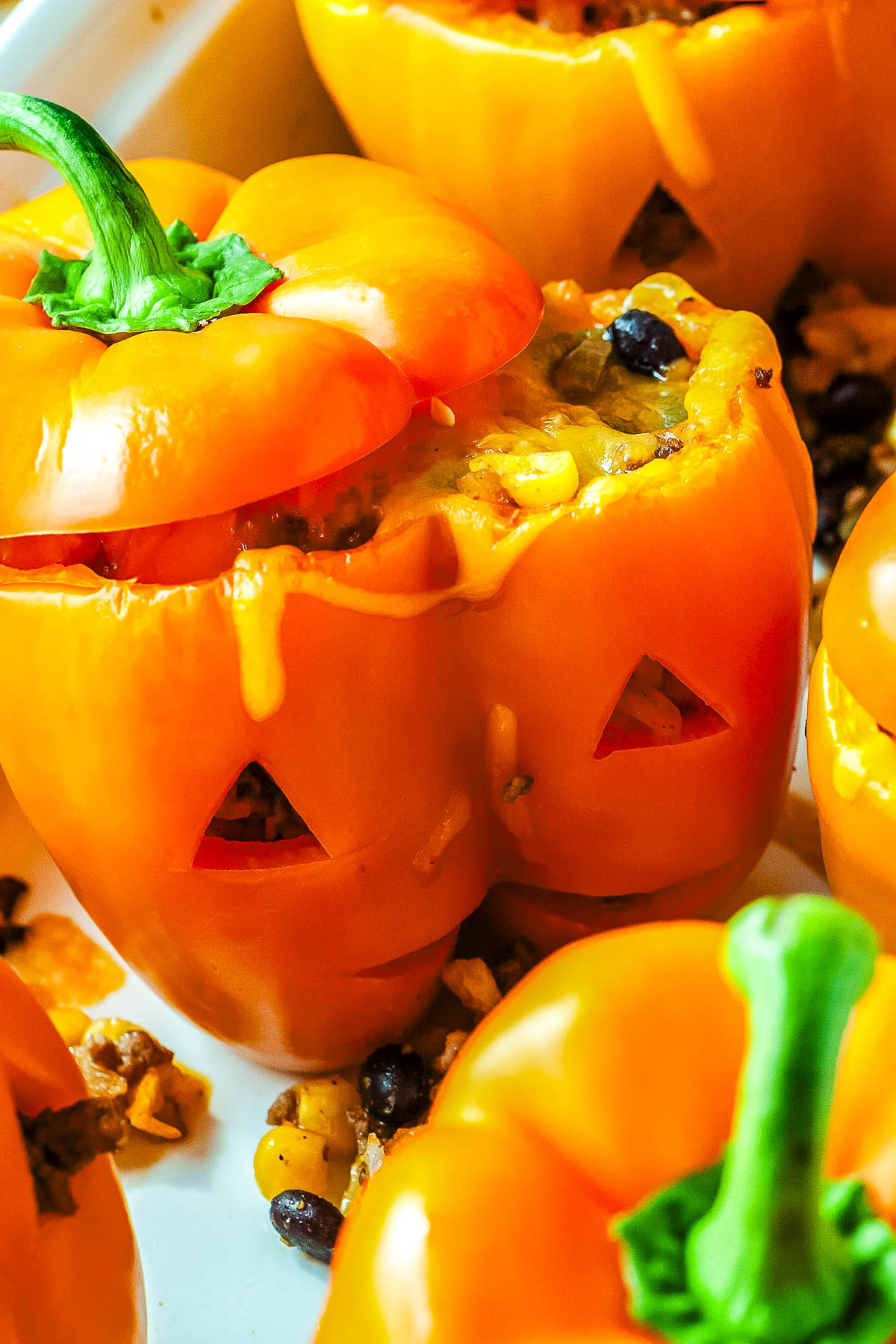A close-up view of a baked orange bell pepper carved with triangular eyes and a jack-o'-lantern grin, filled with seasoned ground beef, corn, black beans, and melted cheddar under a lifted pepper stem cap.
