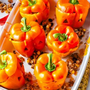 A vertical shot of a white rectangular baking dish filled with six tender baked orange bell peppers carved with jack-o'-lantern faces and stuffed with taco meat, black beans, and corn.