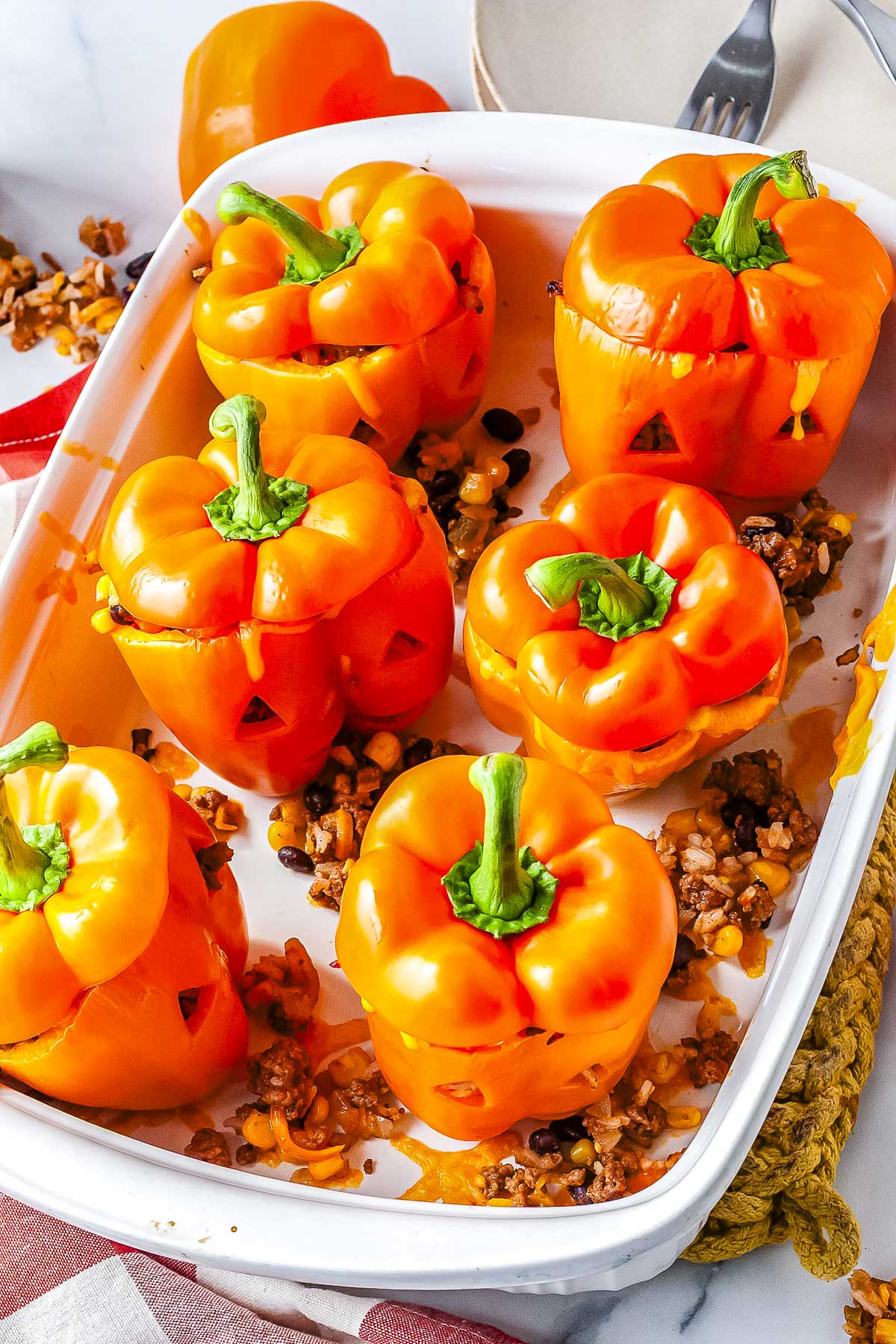 A vertical shot of a white rectangular baking dish filled with six tender baked orange bell peppers carved with jack-o'-lantern faces and stuffed with taco meat, black beans, and corn.