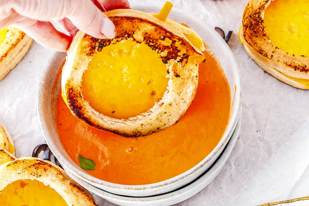 A hand dipping a crisp pumpkin-shaped grilled cheese sandwich with an orange pepper stem into a bowl of creamy tomato soup.