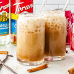 Two can-shaped glasses filled with iced dirty Dr Pepper and sweet cream, styled on a countertop alongside glass straws, Torani syrup bottles, a red Dr Pepper can, and whole cinnamon sticks.