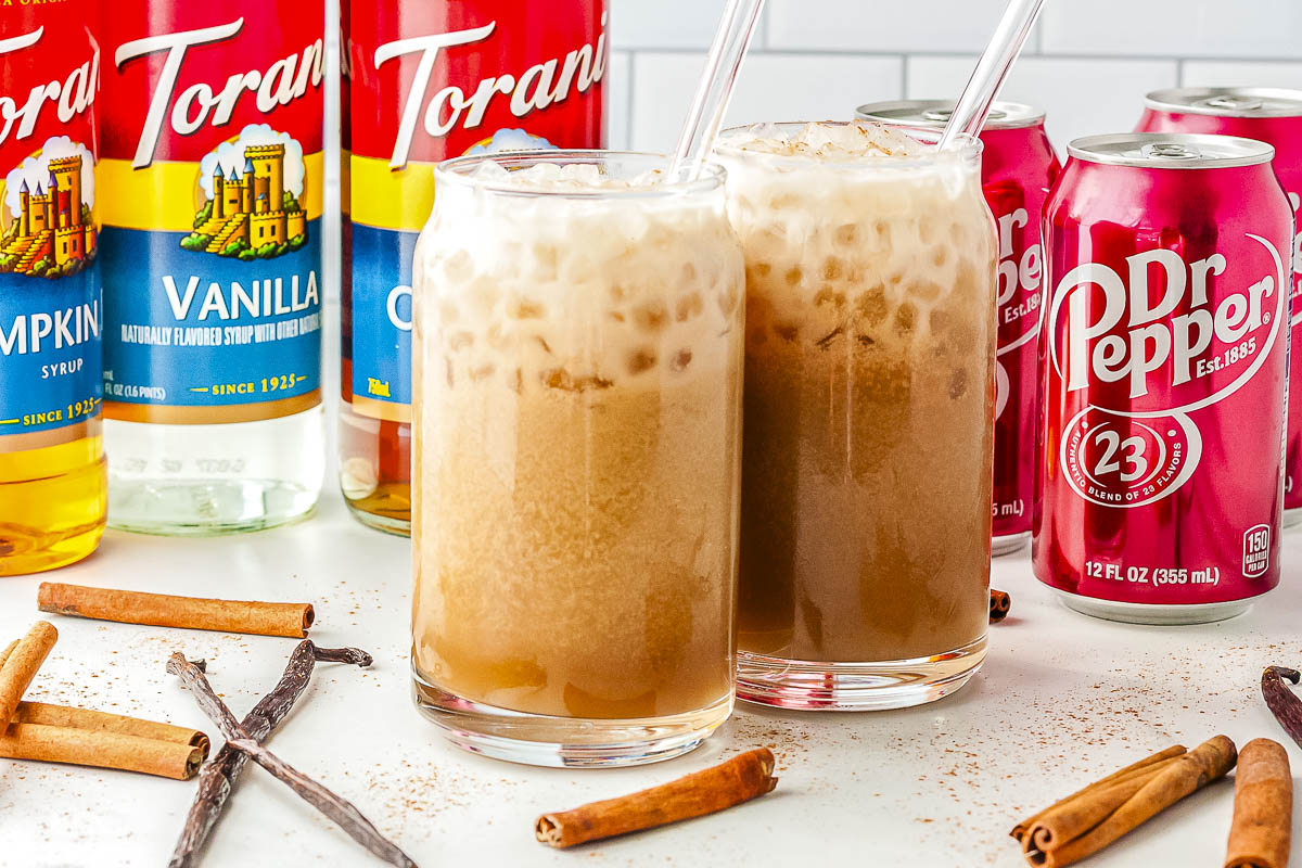 Two can-shaped glasses filled with iced dirty Dr Pepper and sweet cream, styled on a countertop alongside glass straws, Torani syrup bottles, a red Dr Pepper can, and whole cinnamon sticks.
