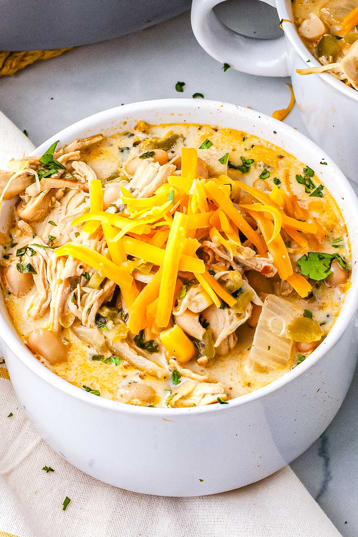 Close view of creamy white chicken chili in a white bowl topped with shredded cheddar cheese, showing shredded chicken, white beans, corn, green chiles, and cilantro.