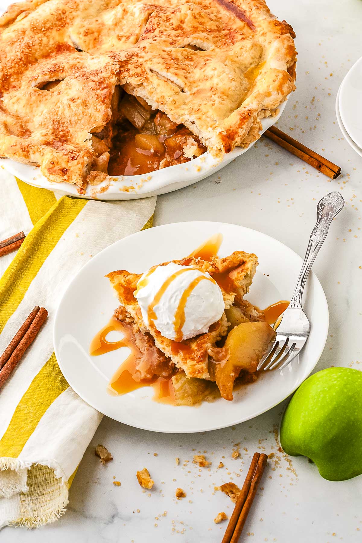 A vertical view of a plated slice of salted caramel apple pie topped with whipped cream and caramel drizzle in front of the whole baked pie dish.