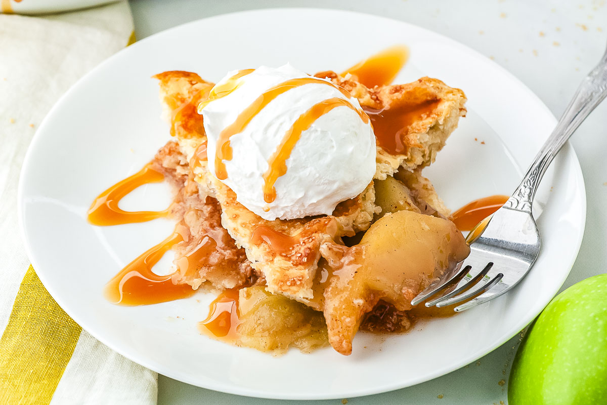 A slice of salted caramel apple pie topped with a scoop of whipped cream and caramel drizzle on a white dessert plate with a silver fork.
