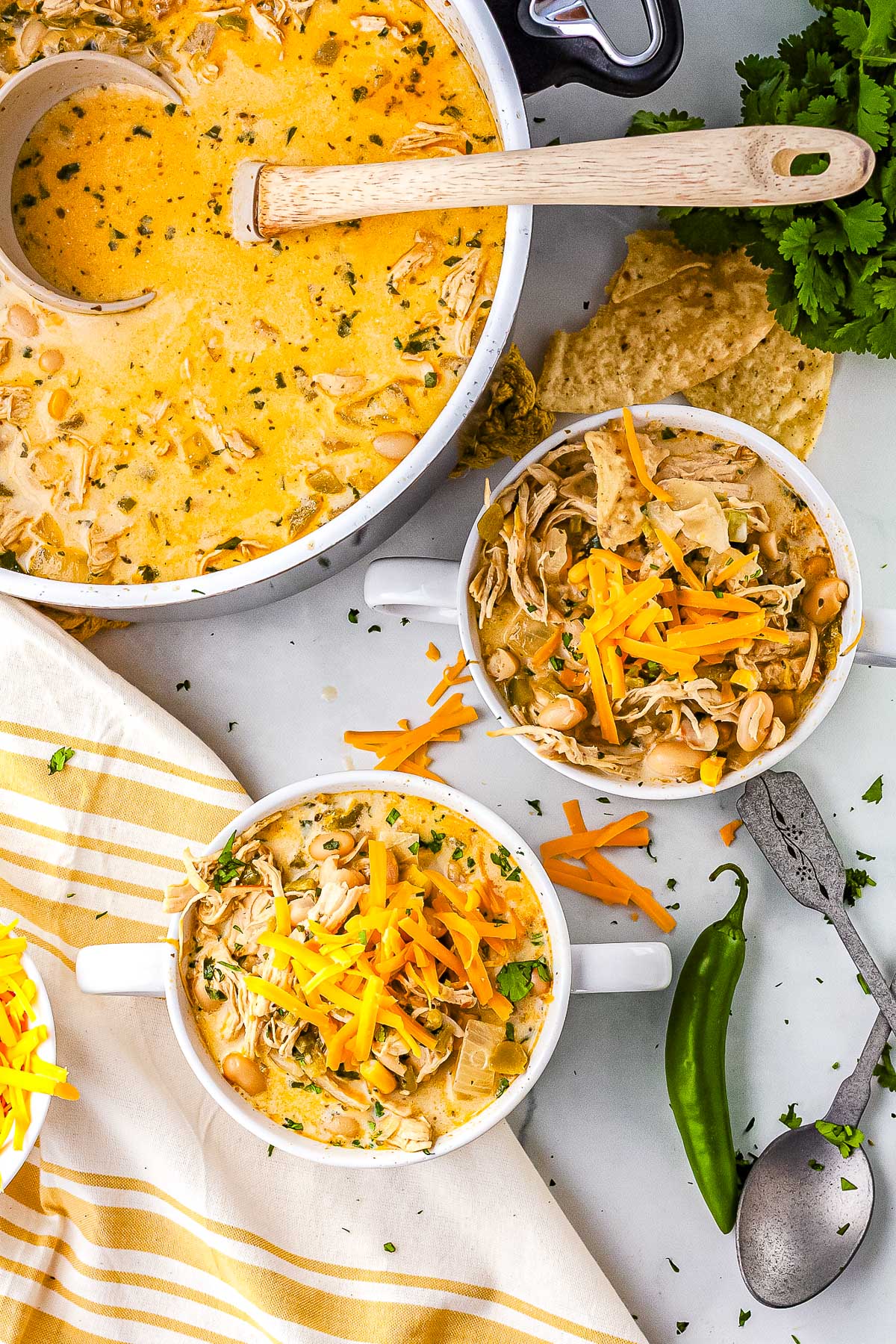Overhead view of homemade white chicken chili in a large pot with two serving bowls topped with shredded cheddar cheese, tortilla chips, and a fresh jalapeño nearby.